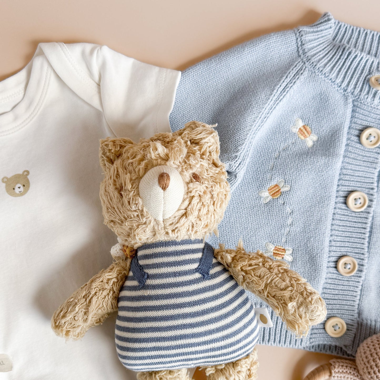 Baby outfit with white onesie, blue cardigan, and teddy bear toy on a beige background