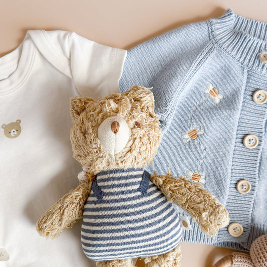 Baby outfit with white onesie, blue cardigan, and teddy bear toy on a beige background