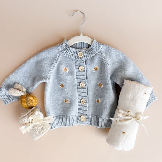 Baby sweater with buttons on a hanger against a beige background, bee lovey and muslin swaddle