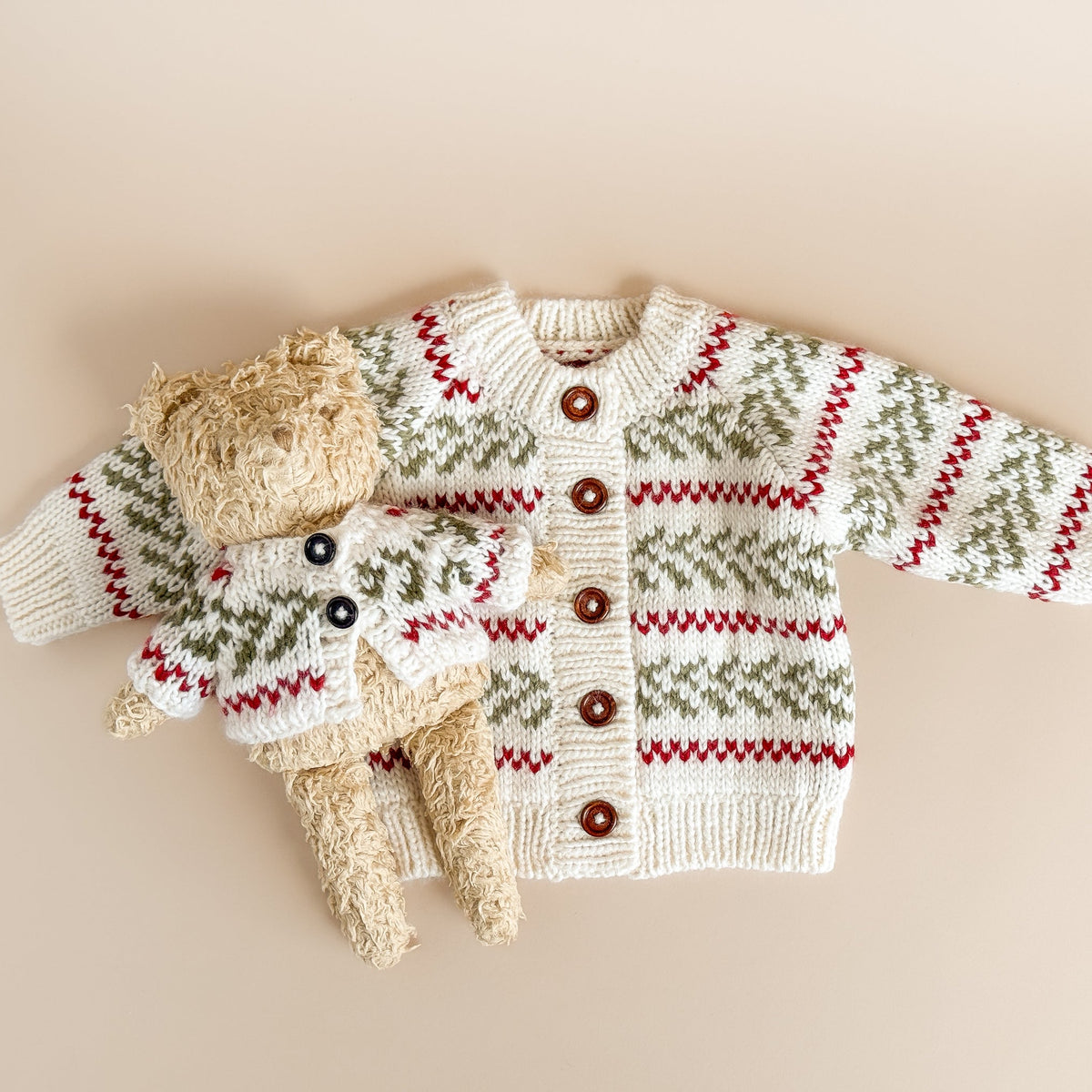 Knitted baby sweater with christmas holiday pattern and buttons on a beige background with teddy bear in matching cardigan