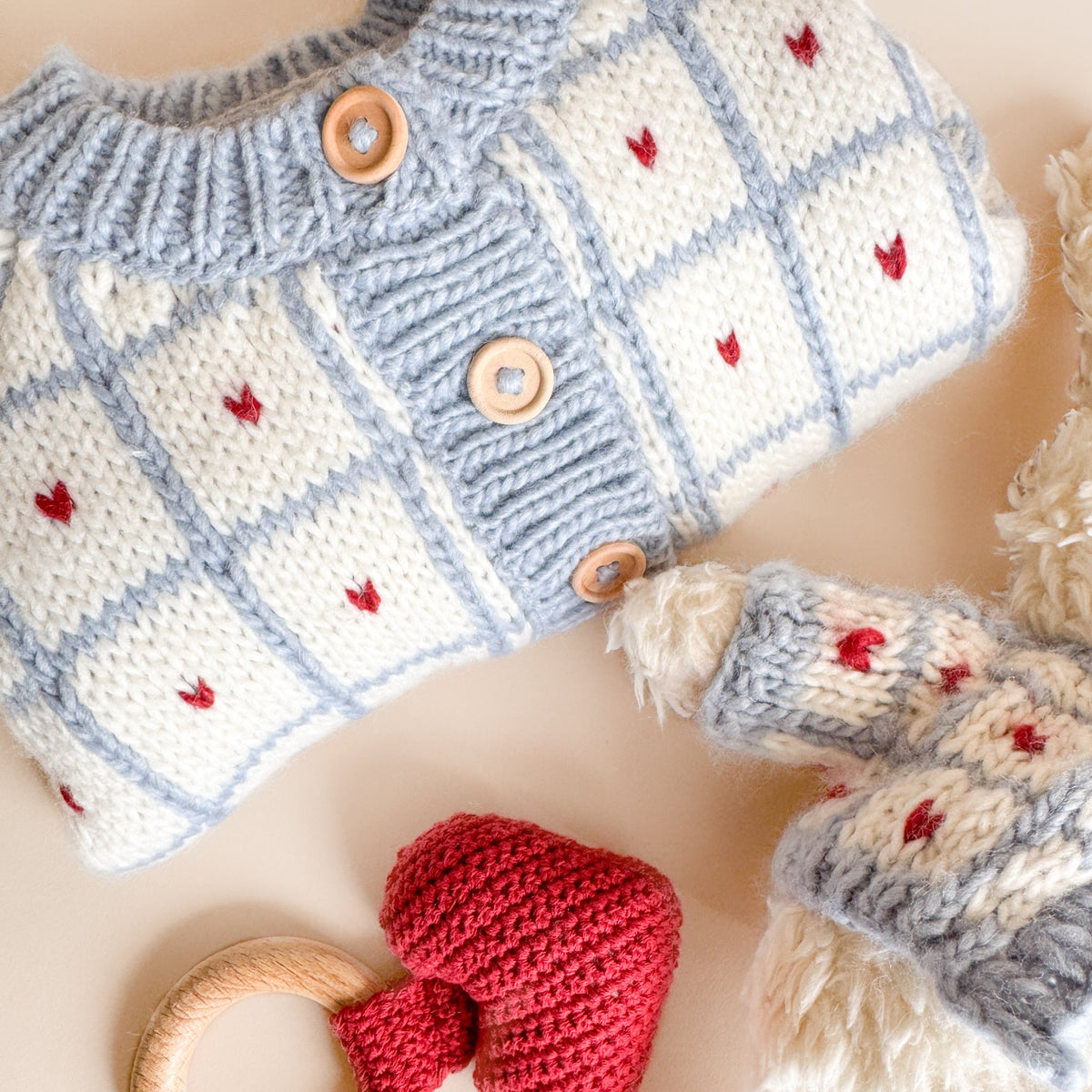 A close-up flat lay of a hand-knit baby cardigan in light blue and cream with small red embroidered motifs and wooden buttons, styled with a red knit heart and wooden ring toy on a warm beige background.